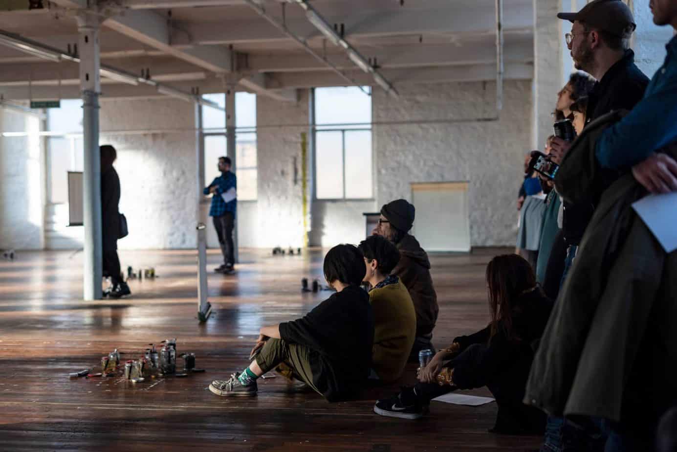 A group of people is gathered in a spacious, well-lit room, with some sitting on the floor and others standing, observing something.