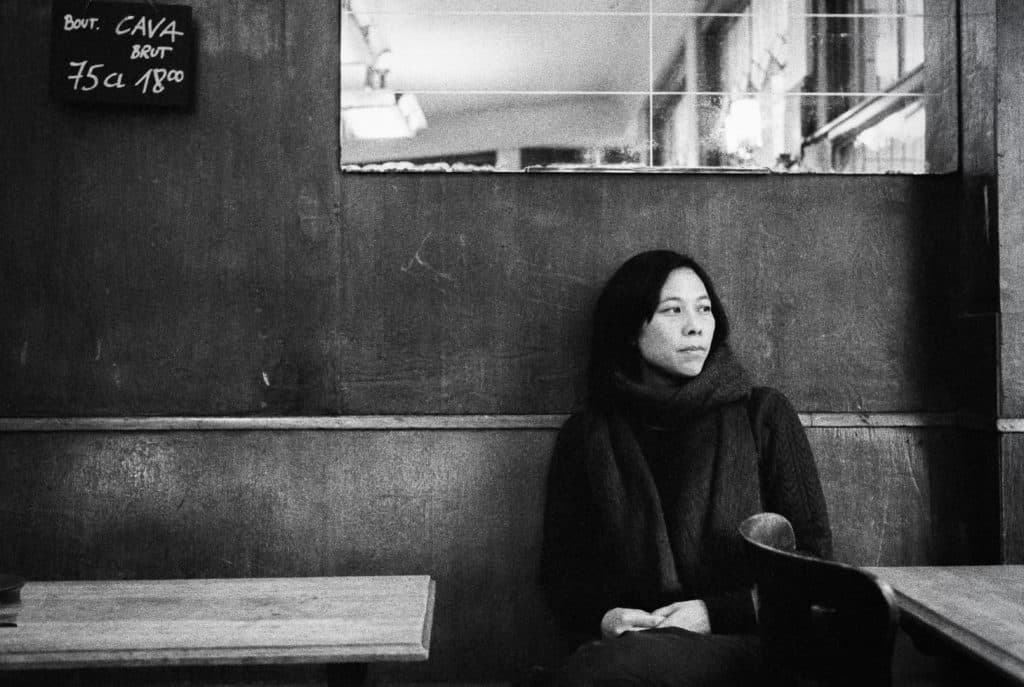 A woman in a scarf sits alone at a table in a dimly lit room with a chalkboard menu on the wall.