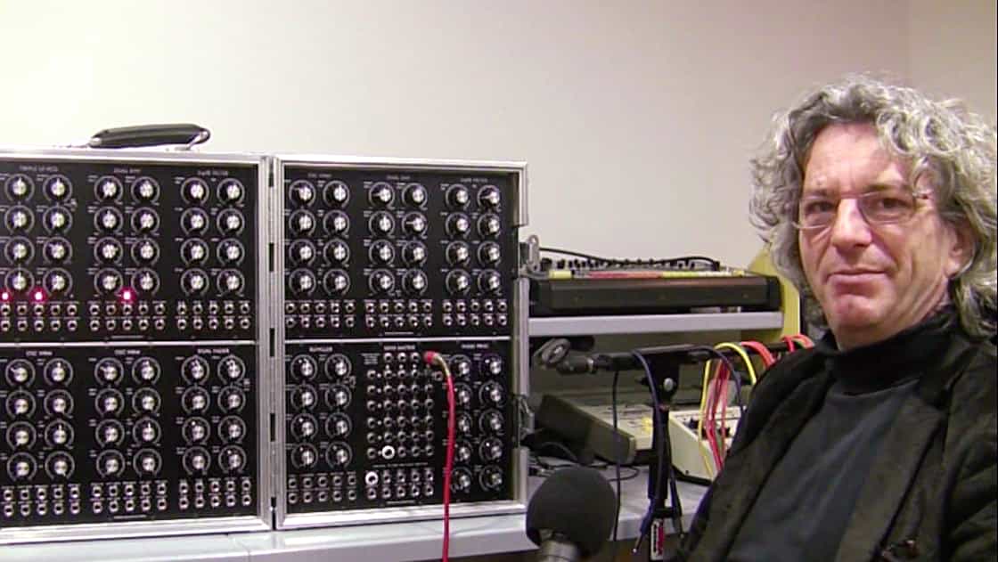 A person with curly hair and glasses sits beside a large modular synthesizer with numerous knobs and cables.