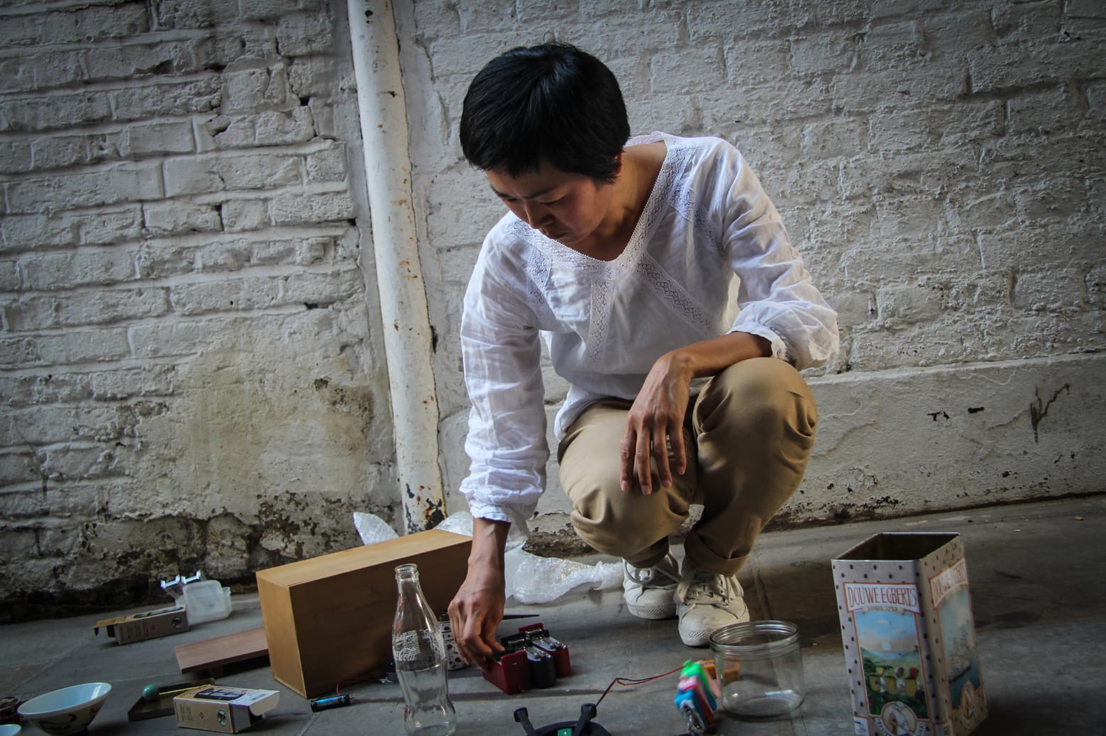 A person in a white shirt and beige pants is crouching on the floor, arranging various items including bottles and boxes near a brick wall.