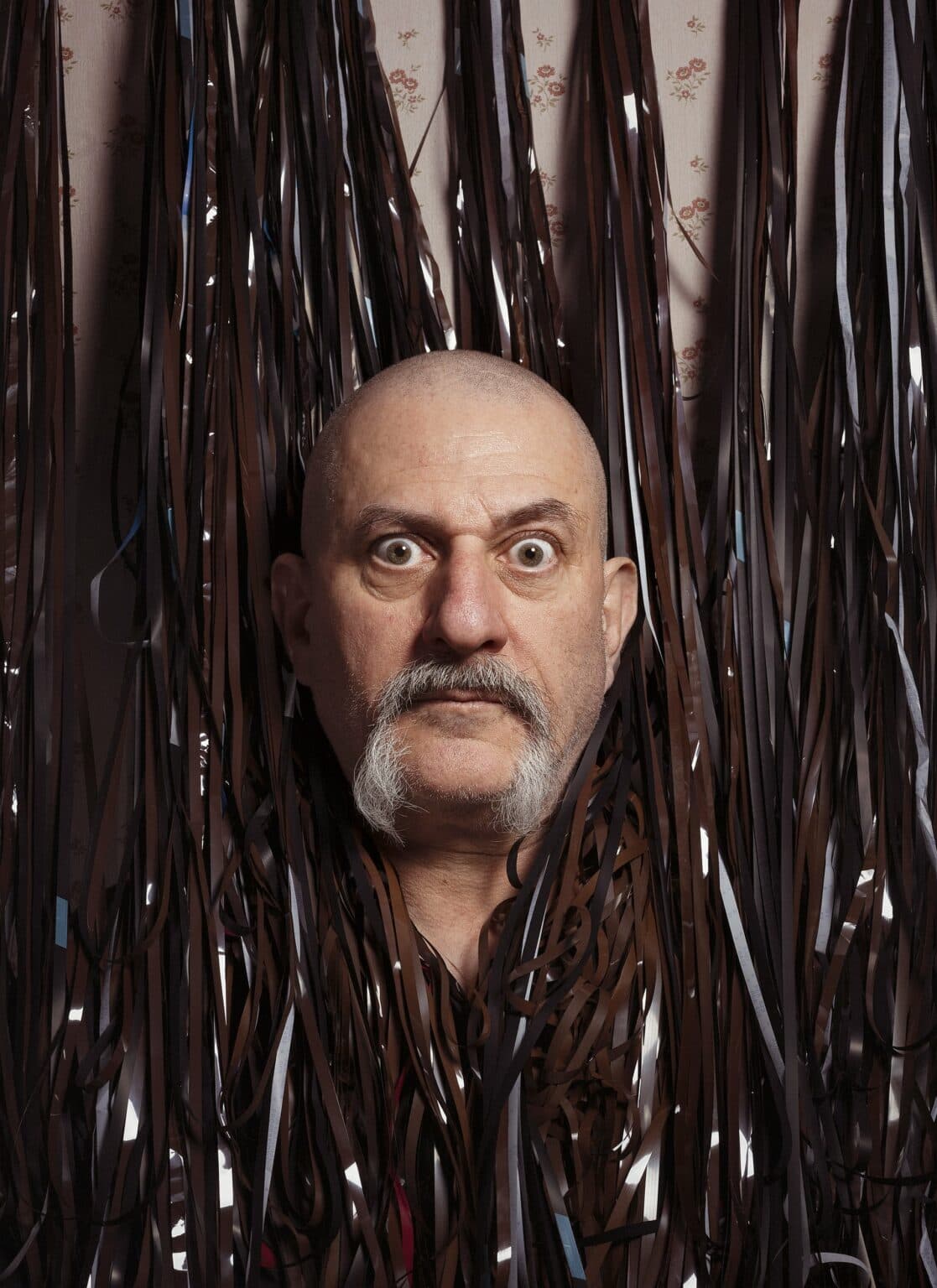 A man with a bald head and a mustache is surrounded by hanging strips of magnetic tape.