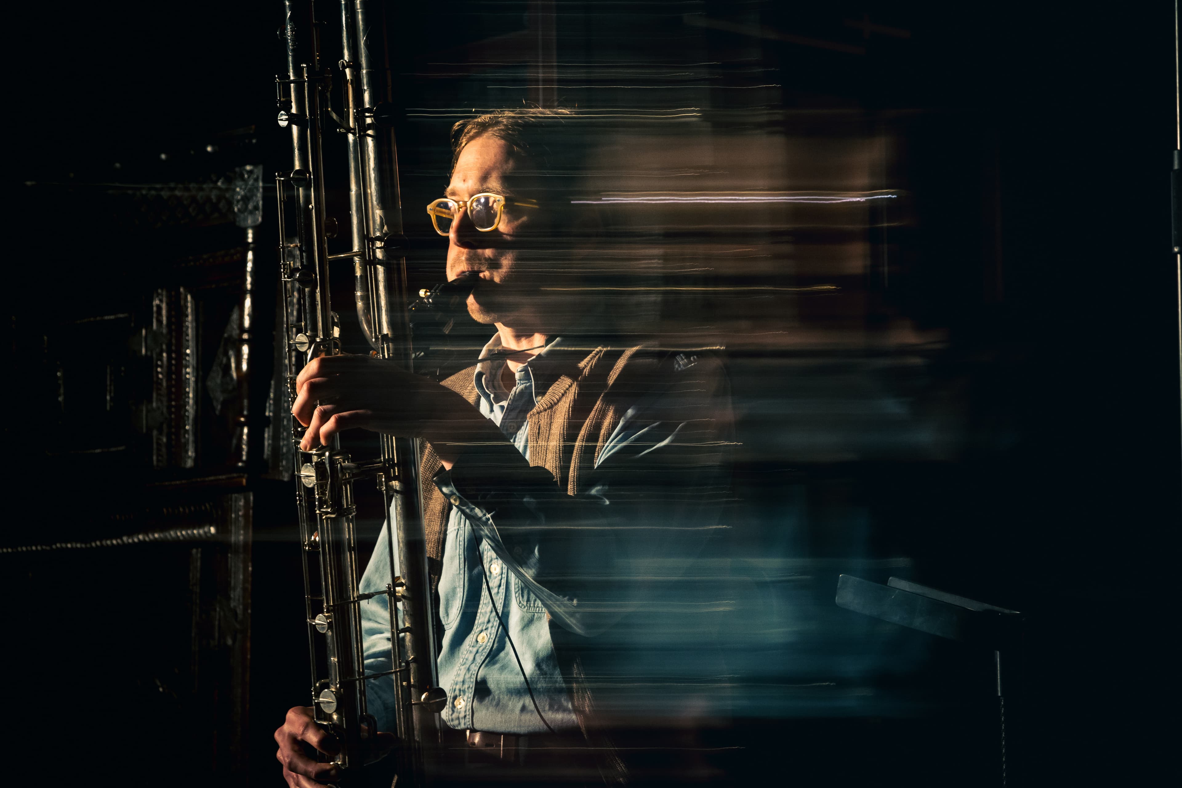 A person wearing glasses plays a contra-bass clarinet with motion blur effects in a dimly lit setting.