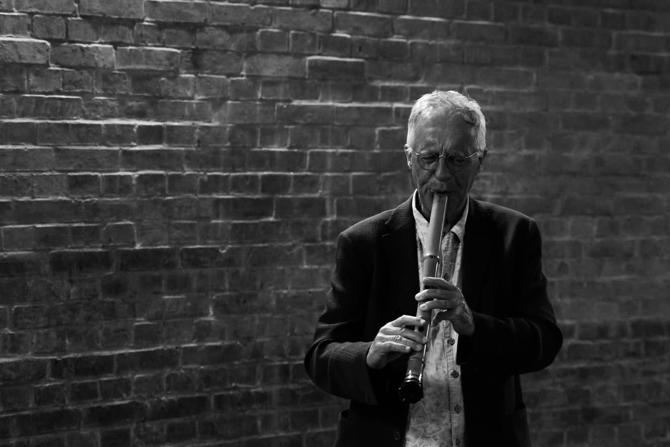 An elderly man plays a shakuhachi flute against a brick wall in a black and white setting.